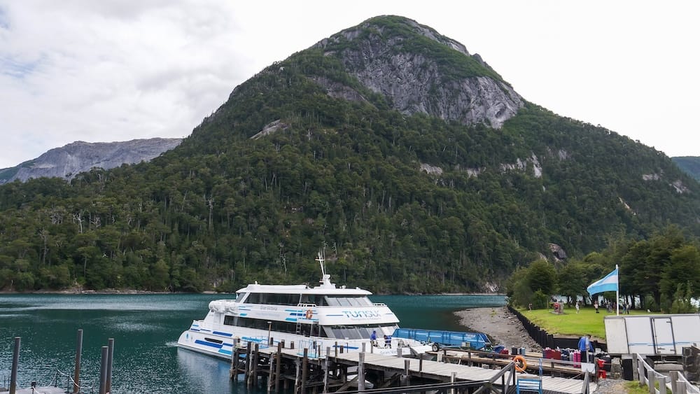 Catamaran at Puerto Blest on Nahuel Huapi Lake beneath Patagonian mountain slopes A white tourist catamaran is moored at Puerto Blest on Nahuel Huapi Lake in Río Negro, Patagonia, Argentina, beneath a steep forested mountain with exposed rock rising directly behind the pier.