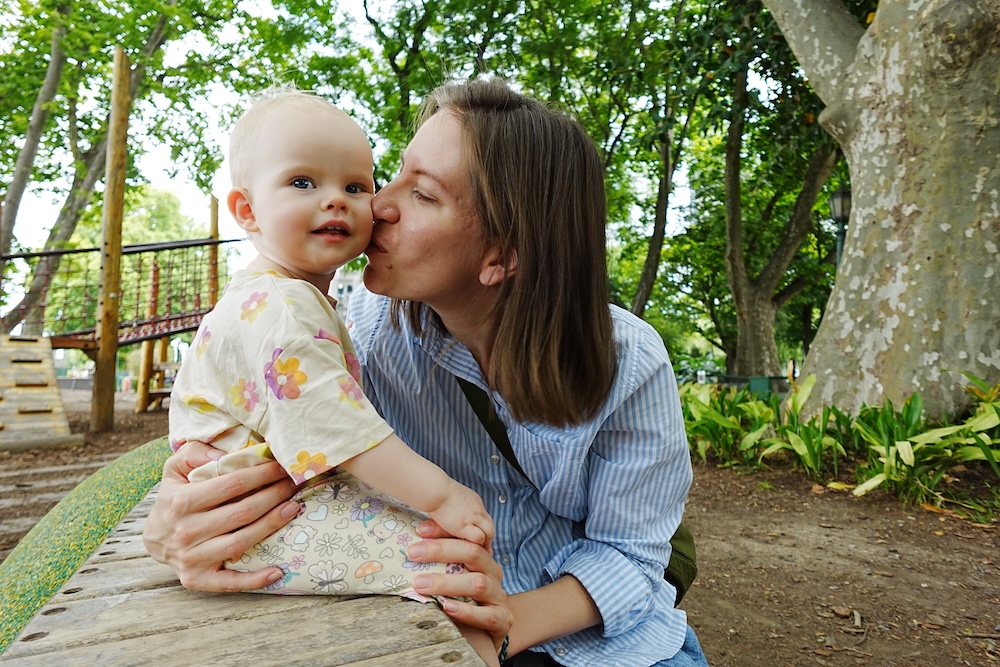Puerto Madero, Buenos Aires, Argentina, where Audrey Bergner kisses her baby Aurelia on a park bench in a leafy playground, capturing a gentle family travel moment while exploring the waterfront district during a relaxed afternoon in the city.