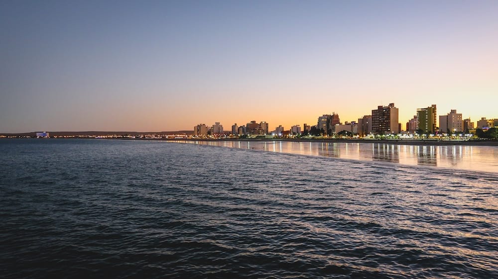 Puerto Madryn’s coastal skyline glows at golden hour in Chubut, Patagonia, Argentina, with apartment towers, waterfront lights and warm reflections stretching across the shallow tidal water.