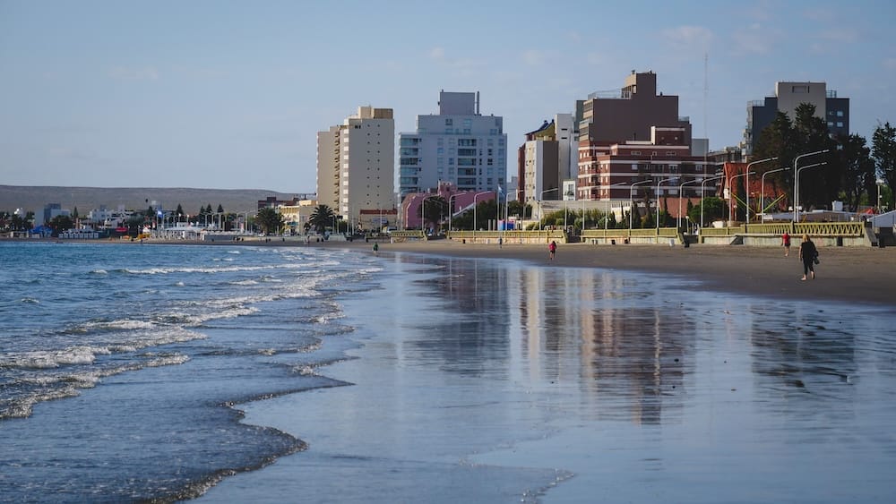Costanera, playa y perfil urbano de Puerto Madryn, Chubut, Patagonia, Argentina, durante una mañana tranquila Pequeñas olas matutinas llegan a la playa de Puerto Madryn, Chubut, Patagonia, Argentina, donde la arena mojada refleja los edificios costeros, el paseo marítimo y algunas personas caminando junto al golfo.