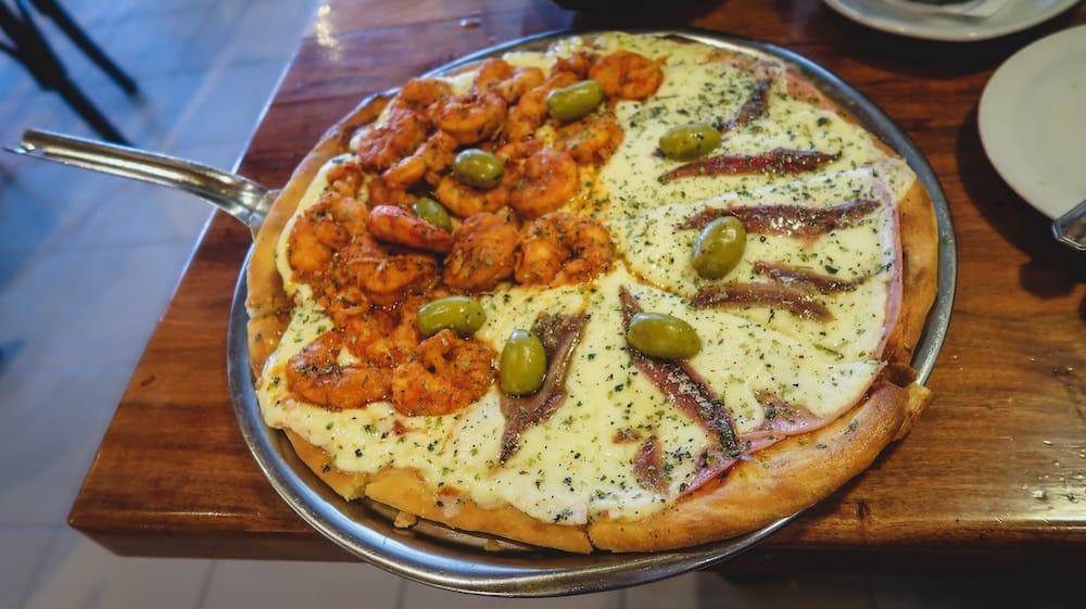 Half shrimp and anchovy seafood pizza served on a metal tray in Puerto Madryn, Argentina, showcasing a budget-friendly Patagonian coastal meal during siesta hours when travelers rely on discounted early dinner deals.