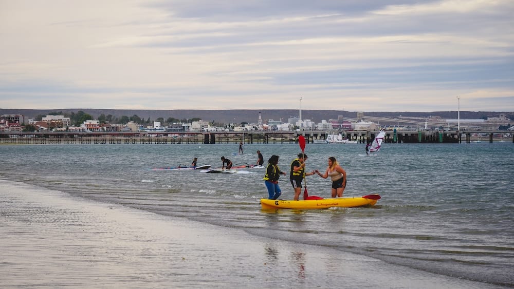 Tres personas preparan un kayak amarillo en aguas poco profundas de la costa de Puerto Madryn, Chubut, Patagonia, Argentina, con practicantes de paddle surf, un muelle largo y la ciudad costera detrás.