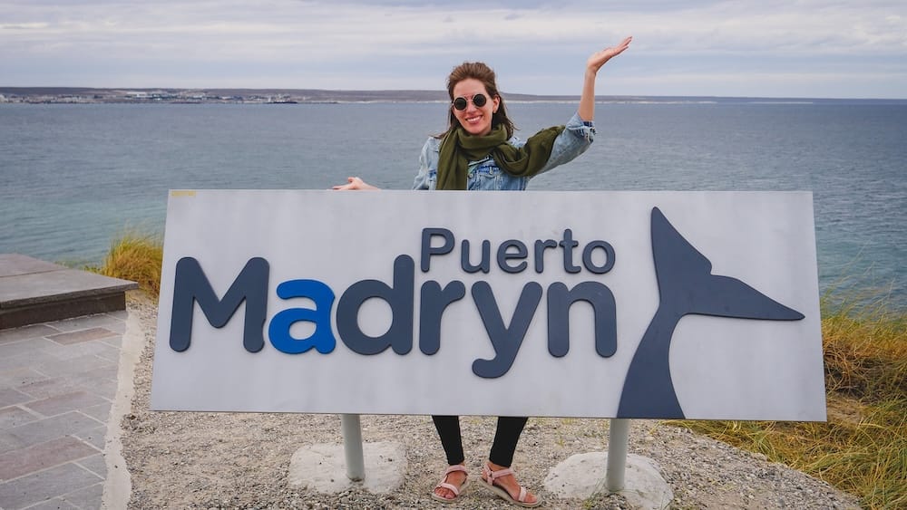 Puerto Madryn Welcome Sign — Chubut, Patagonia, Argentina | Atlantic Coast Photo with Audrey Bergner Audrey Bergner smiles and waves beside the large Puerto Madryn welcome sign, overlooking the Atlantic coastline in Chubut, Argentine Patagonia, with grassy dunes and open ocean in the background.