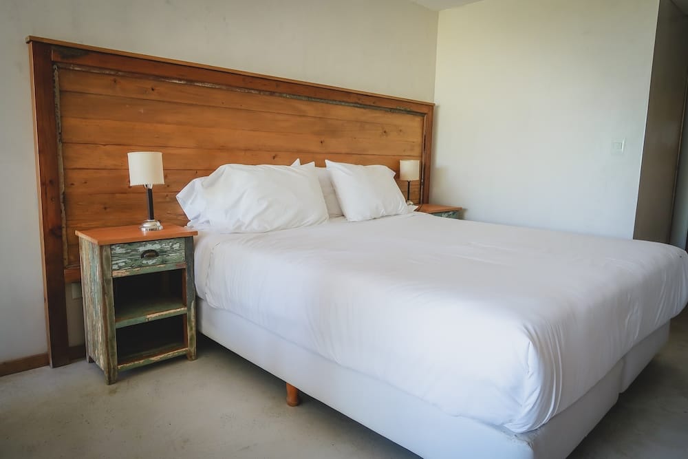 A bright accommodation room in Puerto Pirámides, Chubut, Patagonia, Argentina, with a large white bed, rustic wooden headboard, mismatched bedside tables and simple walls, showing the kind of compact base used for a stay on Península Valdés.
