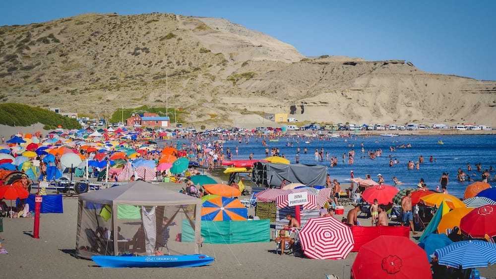 Crowds fill the beach and shallow water at Puerto Pirámides on Península Valdés in Chubut, Patagonia, Argentina, with colourful umbrellas, kayaks, parked vehicles and pale coastal cliffs under a clear blue sky.