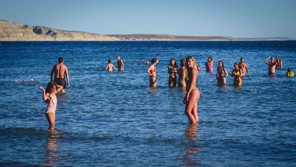 Swimmers cooling off in the blue water at Puerto Pirámides on Península Valdés, Chubut, during a hot summer day in Patagonia, Argentina, with dry coastal cliffs across the bay.
