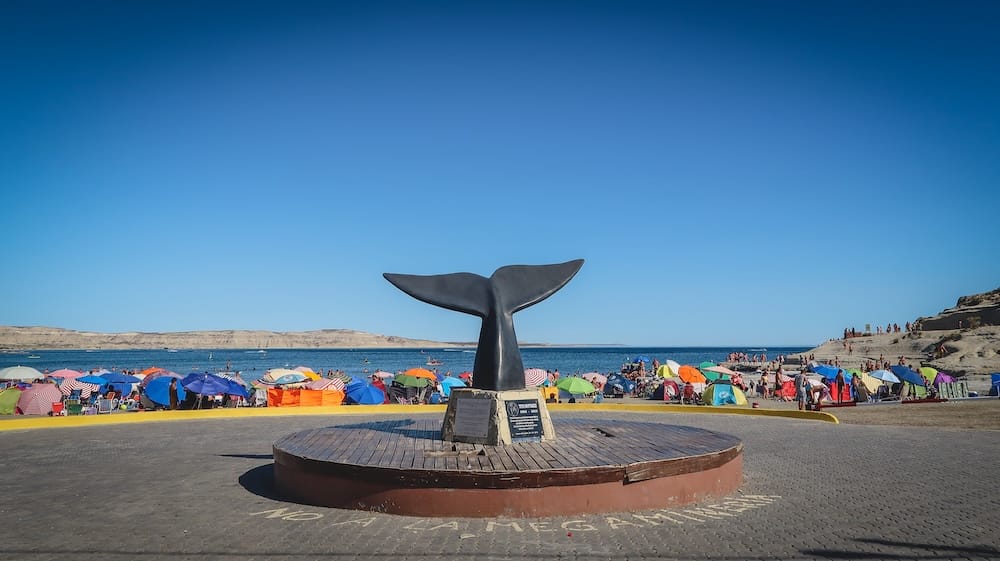 Monumento a la cola de ballena sobre la playa de Puerto Pirámides, Península Valdés, Chubut, Patagonia, Argentina El monumento con forma de cola de ballena se alza sobre la playa de Puerto Pirámides, Península Valdés, Chubut, Patagonia, Argentina, rodeado de sombrillas, bañistas y acantilados costeros bajos.