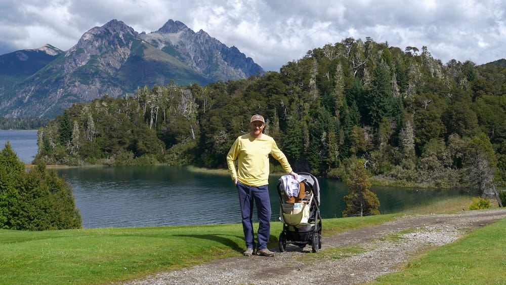 Samuel and Aurelia Jeffery on the walk to Puerto Spiegel near Llao Llao Hotel, Patagonia, Argentina Samuel Jeffery stands beside Aurelia Jeffery in a stroller on the walk toward Puerto Spiegel near Llao Llao Hotel in Bariloche, Río Negro, Patagonia, Argentina, with calm water, forest and jagged mountain peaks behind them.