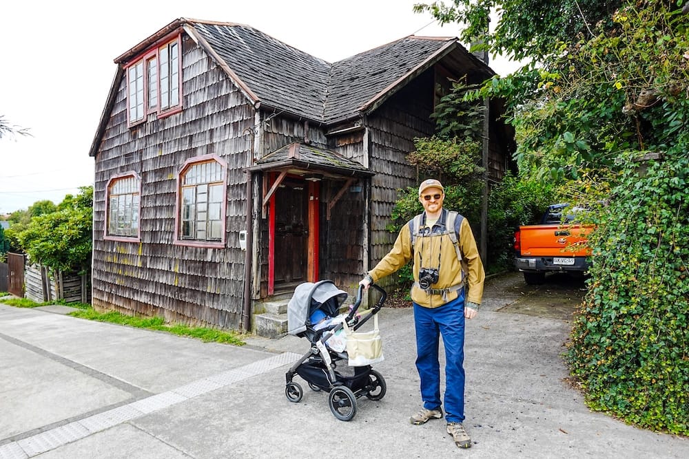 Puerto Varas Family Travel With Stroller and Rain Gear in Los Lagos Chile With Samuel Jeffery and Aurelia Jeffery Samuel Jeffery walking with Aurelia Jeffery in a stroller beside traditional wooden architecture in Puerto Varas, Chile, showing how Patagonia family travel depends on season, rain gear, smooth paths and open town services.