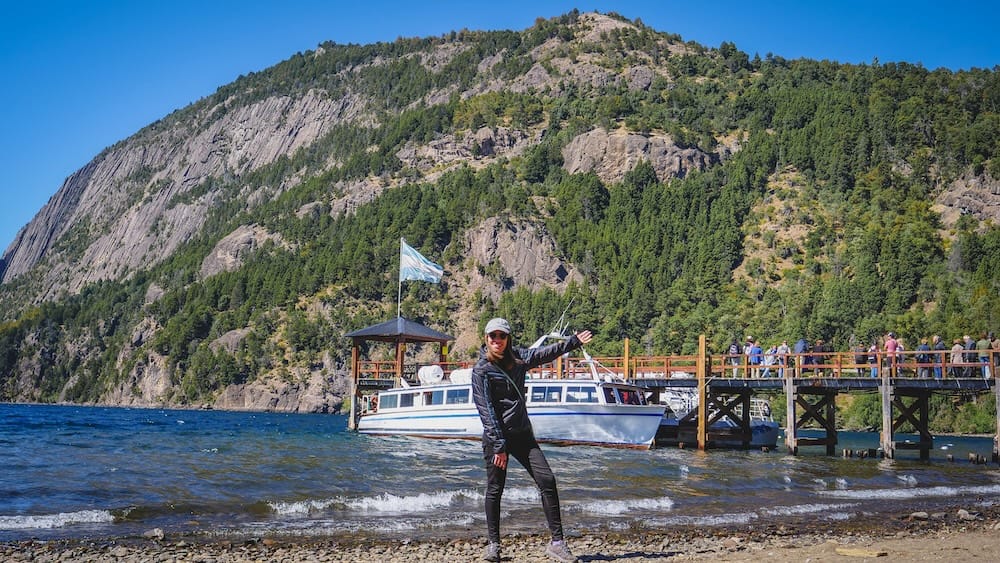Audrey Bergner stands on the rocky shore at Quila Quina Pier on Lake Lácar near San Martín de los Andes, Neuquén, Patagonia, Argentina, with an excursion boat, wooden dock and steep forested mountains behind her.