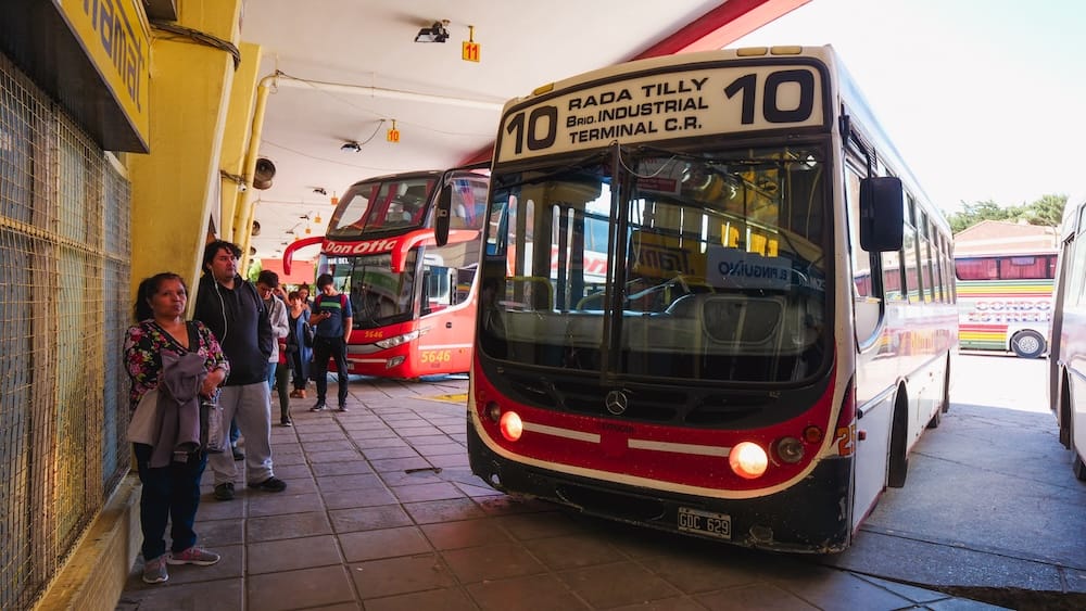 Rada Tilly route 10 bus terminal, Chubut Province, Argentine Patagonia — local and long-distance transit Commuters wait on a covered platform beside local bus route 10 to Rada Tilly at a bus terminal in Chubut Province, Argentine Patagonia, with long-distance coaches parked alongside.