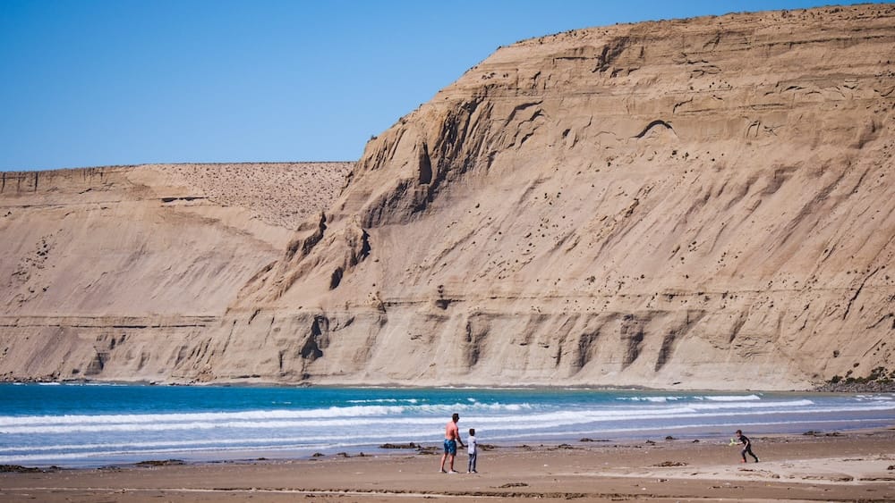 Family walking along the beach in Rada Tilly, Argentina, beneath towering coastal cliffs shaped by erosion, illustrating Patagonia’s wind-exposed Atlantic shoreline and dramatic geological formations.