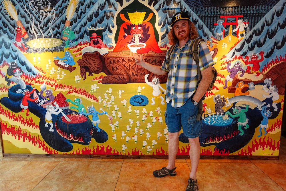 Colourful ramen mural of demons eating noodles in Noboribetsu Onsen, Hokkaido, Japan – Samuel Jeffery Samuel Jeffery, in a cap, plaid shirt and hiking sandals, points at a colourful mural of horned demons cooking and slurping ramen around a crowned figure, inside a building in Noboribetsu Onsen, Hokkaido, Japan.