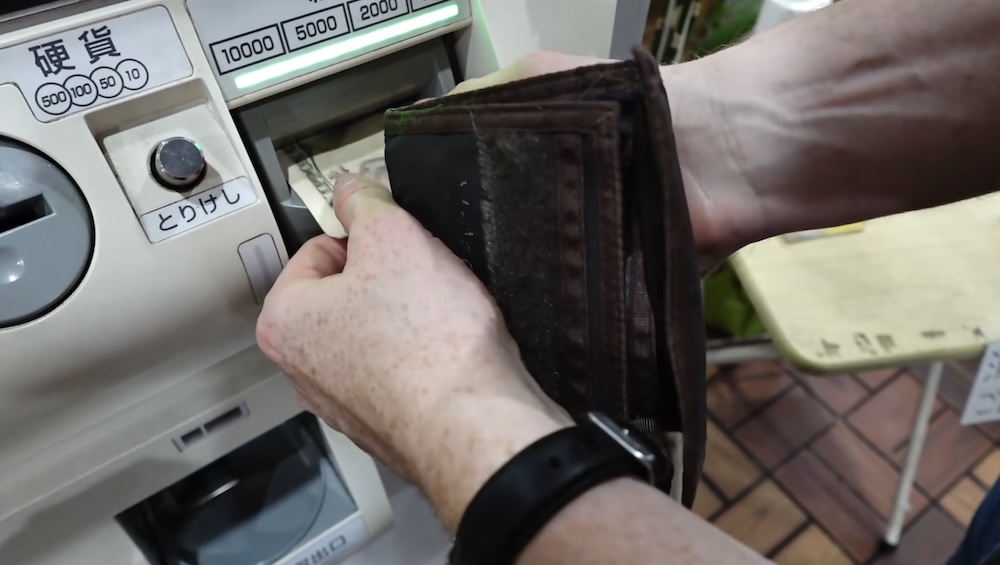 Hands feed a banknote from a brown wallet into the bill slot of a ramen ticket machine in Hakodate, Hokkaido, Japan, beside the coin slot and a cancel button, with the green slot light lit.