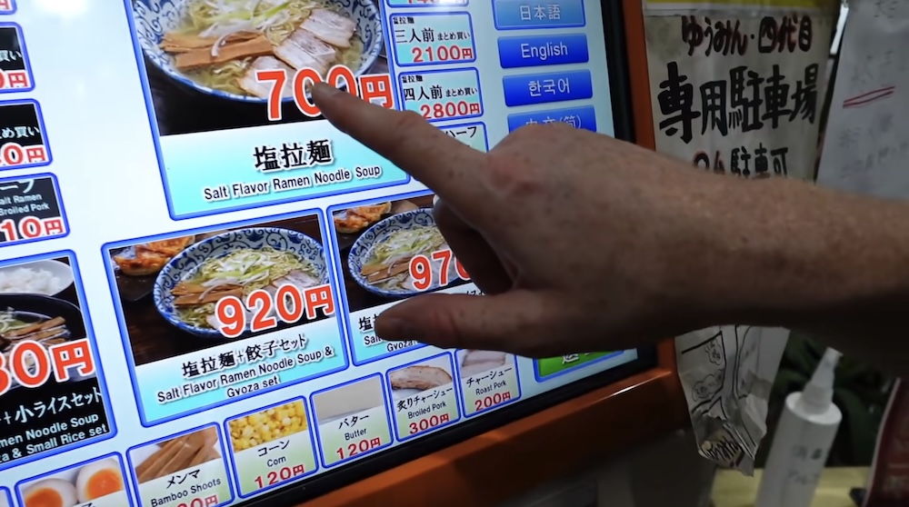 A hand points at the plain salt ramen tile on a ramen ticket machine touchscreen in Hakodate, Hokkaido, Japan, with language buttons including English down the right edge and set tiles and toppings below.