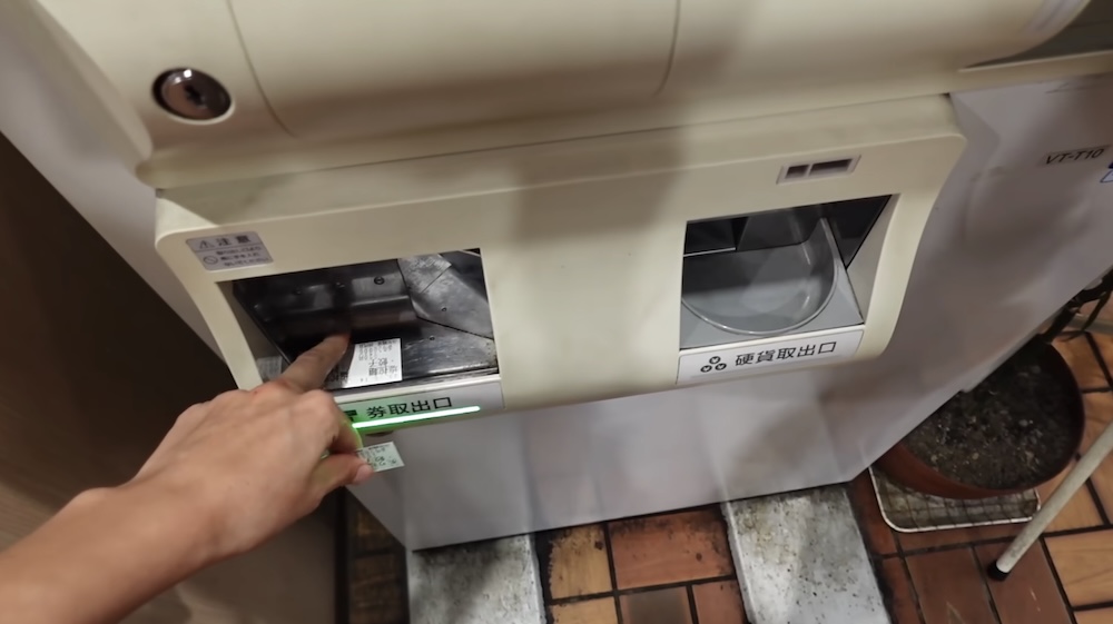 Audrey Bergner’s hand reaches for a printed meal ticket at the ticket outlet of a ramen ticket machine in Hakodate, Hokkaido, Japan, beside a separate coin return tray and a green outlet light.