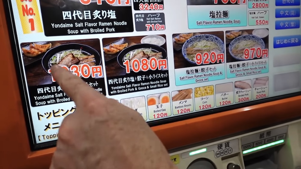 A finger presses the Yondaime salt ramen with broiled pork and gyoza tile on the orange touchscreen of a ramen ticket machine in Hakodate, Hokkaido, Japan, with set tiles, a toppings row and money slots below.