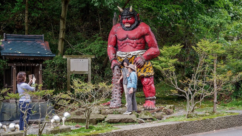 Turistas fotografiando una enorme estatua de oni rojo con un garrote de púas en Noboribetsu Onsen, Hokkaido, Japón Turistas posando para sacarse fotos junto a una enorme estatua de un oni rojo con cuernos, colmillos, barba negra y un gran garrote con púas en Noboribetsu Onsen, Hokkaido, Japón, rodeada de árboles y un pequeño santuario de madera.