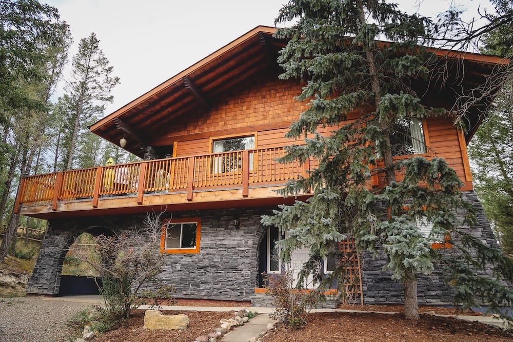 Rental chalet in Invermere, British Columbia, near Kootenay National Park A wood-and-stone chalet with a wraparound deck, rented as accommodation in Invermere, British Columbia, near Radium Hot Springs and Kootenay National Park, surrounded by evergreen trees.