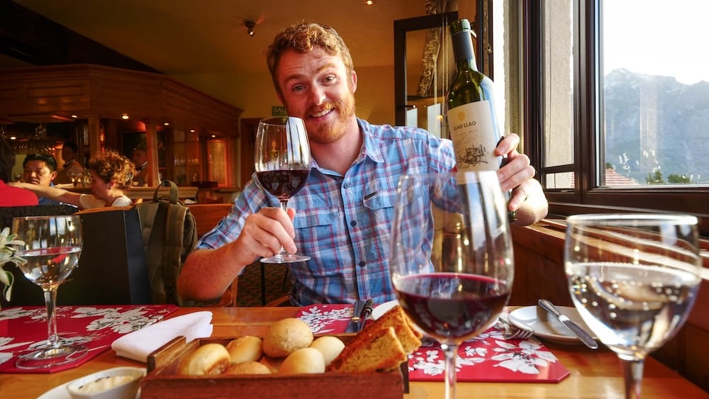 Samuel Jeffery Enjoying Llao Llao Malbec at Restaurante Patagonia in Bariloche, Río Negro, Patagonia, Argentina Samuel Jeffery raises a glass and bottle of Llao Llao Malbec while dining at Restaurante Patagonia in Bariloche, Río Negro, Patagonia, Argentina, with bread on the table and the Andes visible through the window.