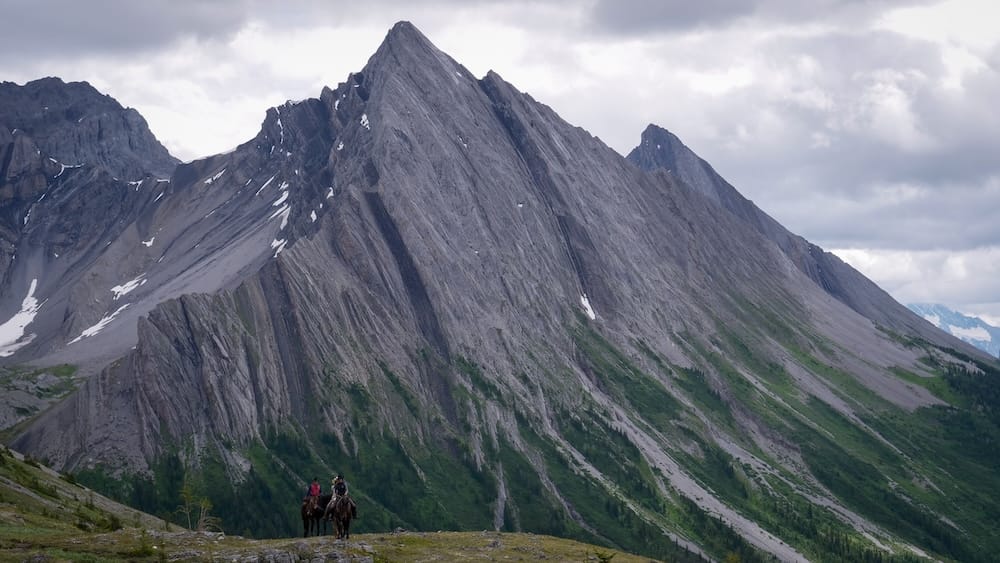 Jinetes empequeñecidos por el pico, Allenby Pass, Banff Trail Riders, Parque Nacional Banff, Alberta, Canadá Dos pequeños jinetes a caballo quedan empequeñecidos por un enorme pico irregular cerca de Allenby Pass en el Parque Nacional Banff, Alberta, Canadá, en una excursión a caballo por el interior con Banff Trail Riders.