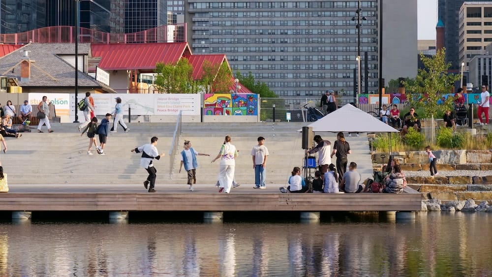 Riverside plaza and pathway signage, Prince's Island Park, Calgary, Alberta, Canada People gather on riverside steps and a wooden dock in Prince's Island Park in Calgary, Alberta, Canada, watching dancers perform near a Peace Bridge wayfinding sign, with downtown high-rises behind them.