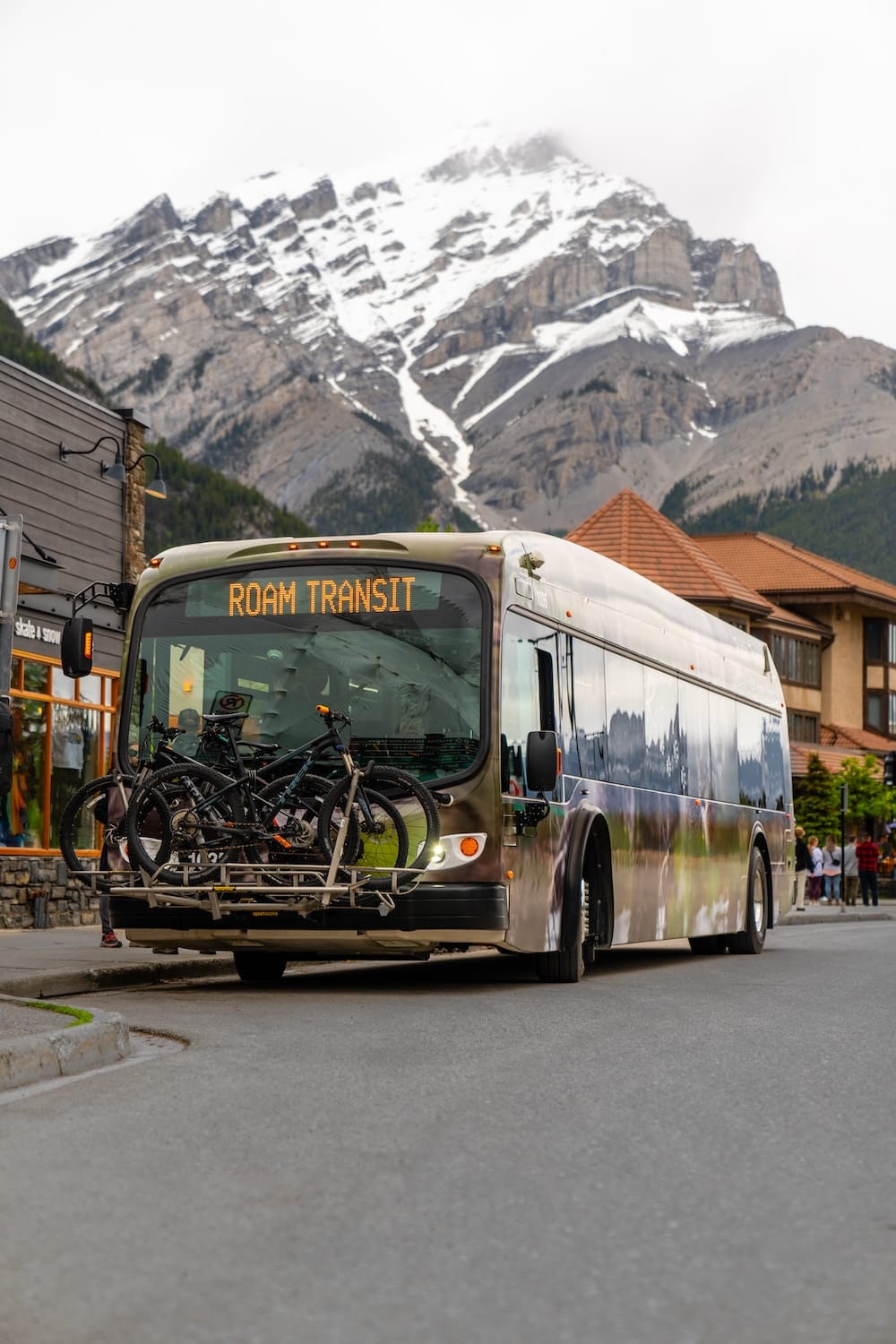 A Roam Transit bus with bicycles mounted on its front rack departs from downtown Banff, Alberta, Canada, with a snow-capped mountain rising directly behind the townsite.