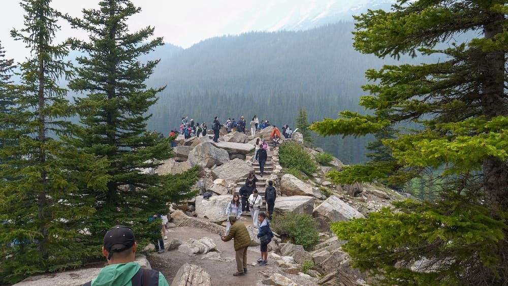 A dense crowd of visitors gathers along the rocky summit of the Rockpile Trail at Moraine Lake in Banff National Park, Alberta, Canada, with forested mountainsides in the hazy background.