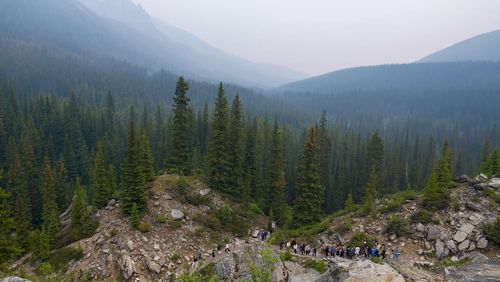 A long line of hikers stretches along the base of the Rockpile Trail at Moraine Lake in Banff National Park, Alberta, Canada, with a forested valley and hazy mountains filling the background.
