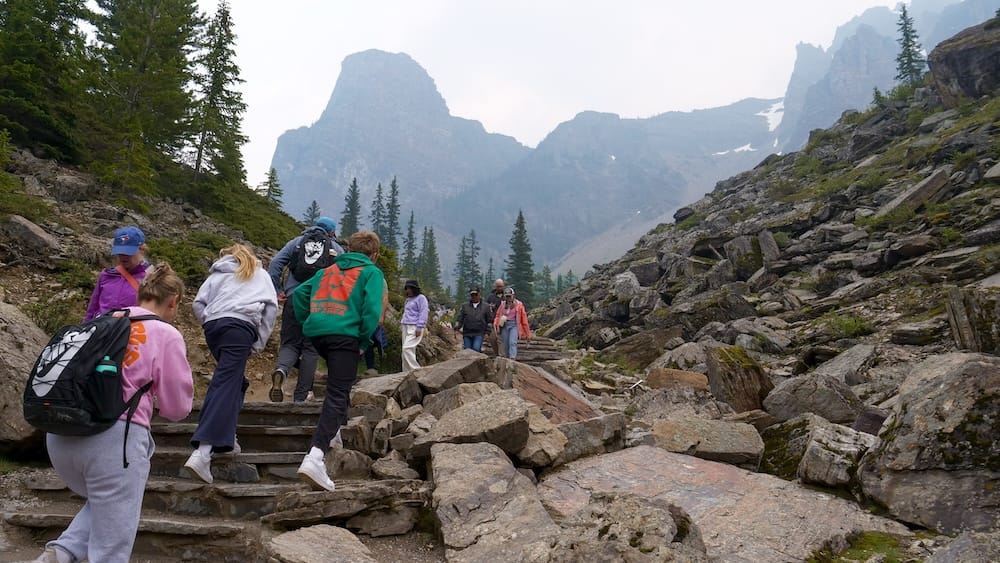 Hikers climb the built stone steps of the Rockpile Trail at Moraine Lake in Banff National Park, Alberta, Canada, surrounded by moraine boulders with mountain peaks visible ahead.