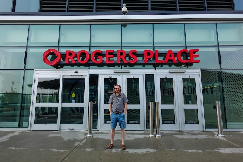 Samuel Jeffery stands outside the main Rogers Place entrance in Edmonton’s ICE District, with the arena’s red signage, glass facade and entry doors visible behind him in Alberta, Canada.