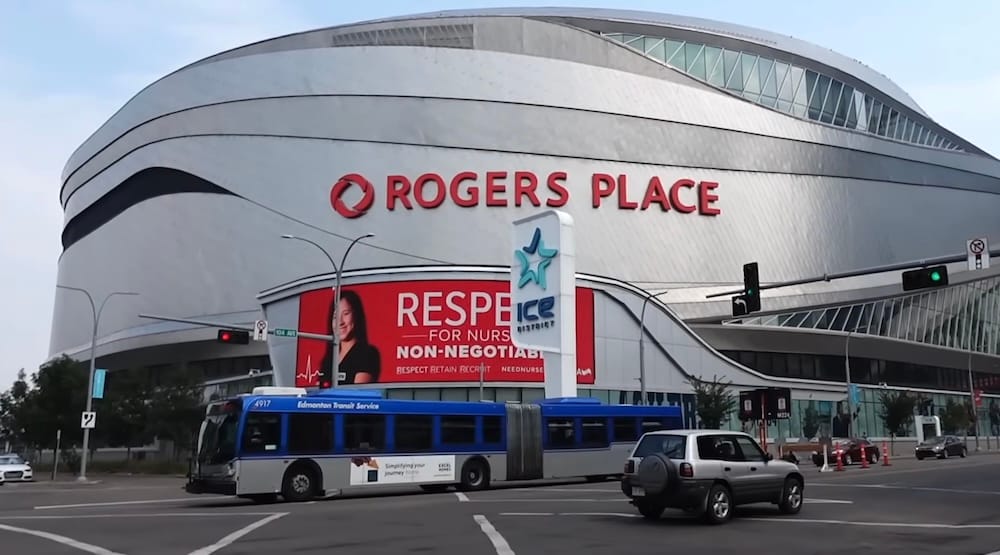 Colectivo articulado de ETS frente a Rogers Place, en el ICE District de Edmonton, Alberta, Canadá Un colectivo articulado del Edmonton Transit Service pasa frente a Rogers Place, en el ICE District de Edmonton, Alberta, Canadá, con el cartel del estadio, el tránsito y la fachada curva plateada detrás.