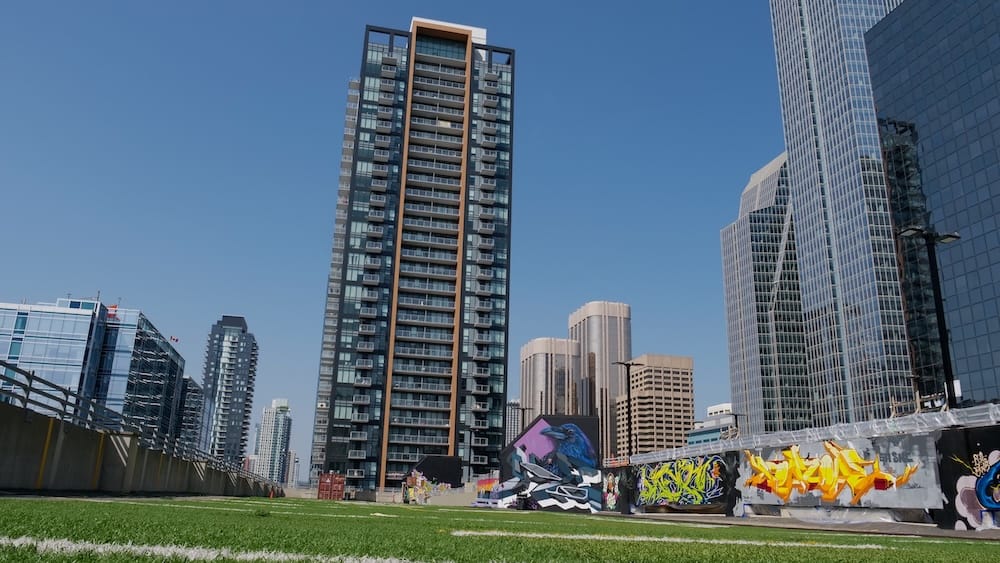 El área con pasto sintético de High Park en Calgary, Alberta, Canadá, bordeada por murales de estilo graffiti, con las torres altas del centro y un cielo despejado de fondo.