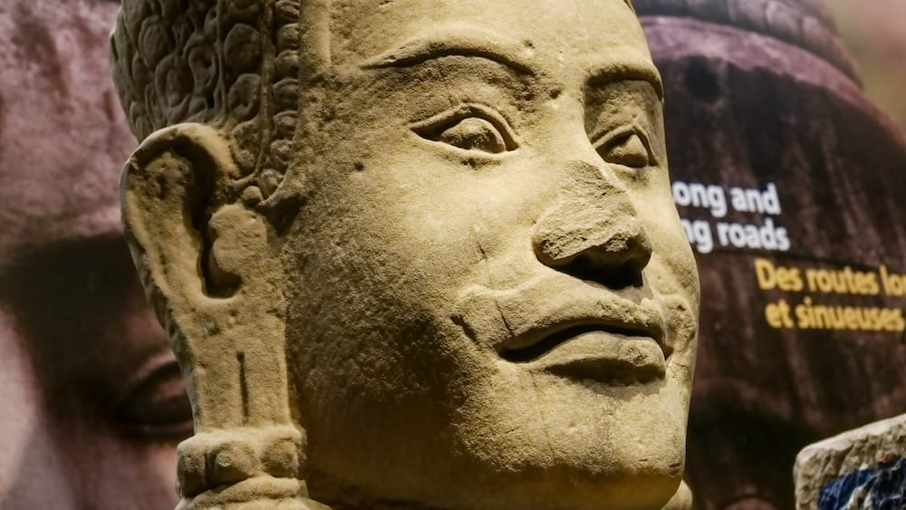 Close view of an ancient carved stone head displayed inside Royal Alberta Museum in Edmonton, Alberta, Canada, with bilingual exhibition text and another large sculpted face visible behind.