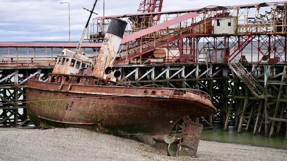 Abandoned Tugboat at the Old Port Pier, Río Gallegos, Santa Cruz Province, Patagonia, Argentina A rusted, abandoned tugboat sits beached on a gravel shore beneath the old industrial loading pier in Río Gallegos, Santa Cruz Province, Argentine Patagonia, its hull streaked with rust and moss below a weathered crane structure.