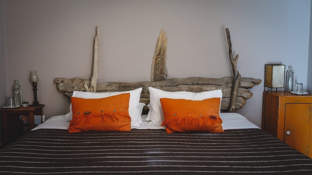 Una acogedora habitación de hotel boutique en Bariloche tiene una cabecera rústica de madera flotante, almohadones tejidos naranjas, ropa de cama blanca y muebles cálidos de madera en Río Negro, Patagonia argentina.