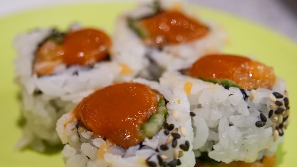 Primer plano de rollos de sushi cubiertos con fetas de salmón y una mezcla de semillas de sésamo blancas y negras, con palta a la vista en el interior, sobre un plato verde en Point Sushi en Calgary, Alberta, Canadá.