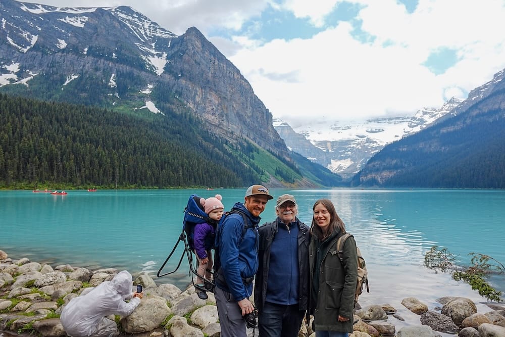 Samuel, Audrey, Daniel y Aurelia en Lake Louise, Parque Nacional Banff, Alberta, Canadá. Samuel Jeffery, Audrey Bergner y Daniel Bergner posan para una foto familiar en Lake Louise en el Parque Nacional Banff, Alberta, Canadá, con la bebé Aurelia Jeffery en una mochila portabebés y otro visitante fotografiando la orilla en primer plano.