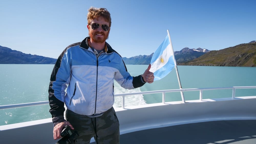 Boat deck on Lago Argentino near El Calafate, Santa Cruz, Patagonia, Argentina Samuel Jeffery smiles with a thumbs up beside an Argentine flag on the deck of a boat crossing the turquoise waters of Lago Argentino near El Calafate, Santa Cruz Province, Patagonia, Argentina.