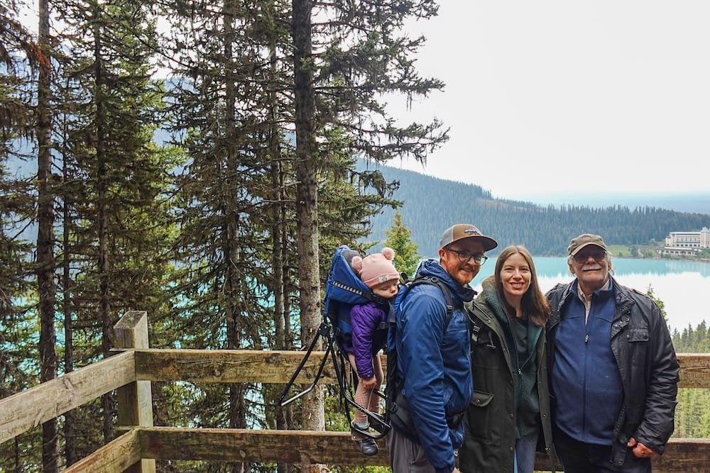 Samuel Jeffery, Audrey Bergner, and Daniel Bergner stand at a wooden viewpoint railing above turquoise Lake Louise with Aurelia in a child carrier, forested slopes and the Fairmont Chateau Lake Louise visible below, Banff National Park, Alberta, Canada.
