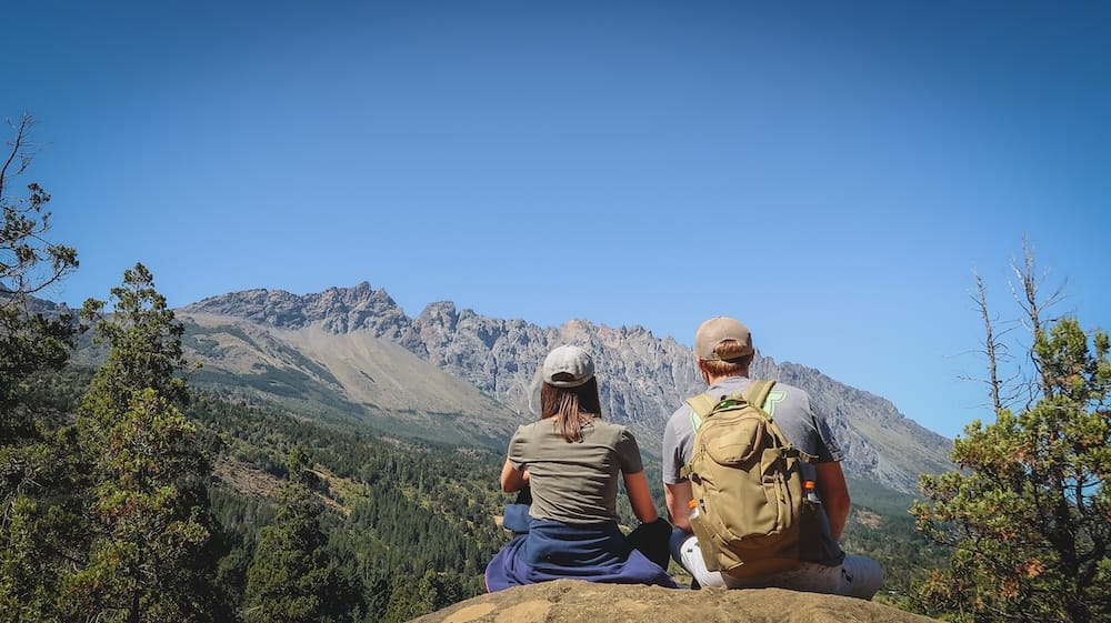 Hikers Overlooking El Bolsón, Río Negro Province, Patagonia, Argentina — Samuel Jeffery and Audrey Bergner Samuel Jeffery and Audrey Bergner sit with their backs to the camera on a rocky outcrop above El Bolsón, Río Negro Province, in Northern Argentine Patagonia, taking in a panoramic view of jagged mountain peaks and forested slopes.