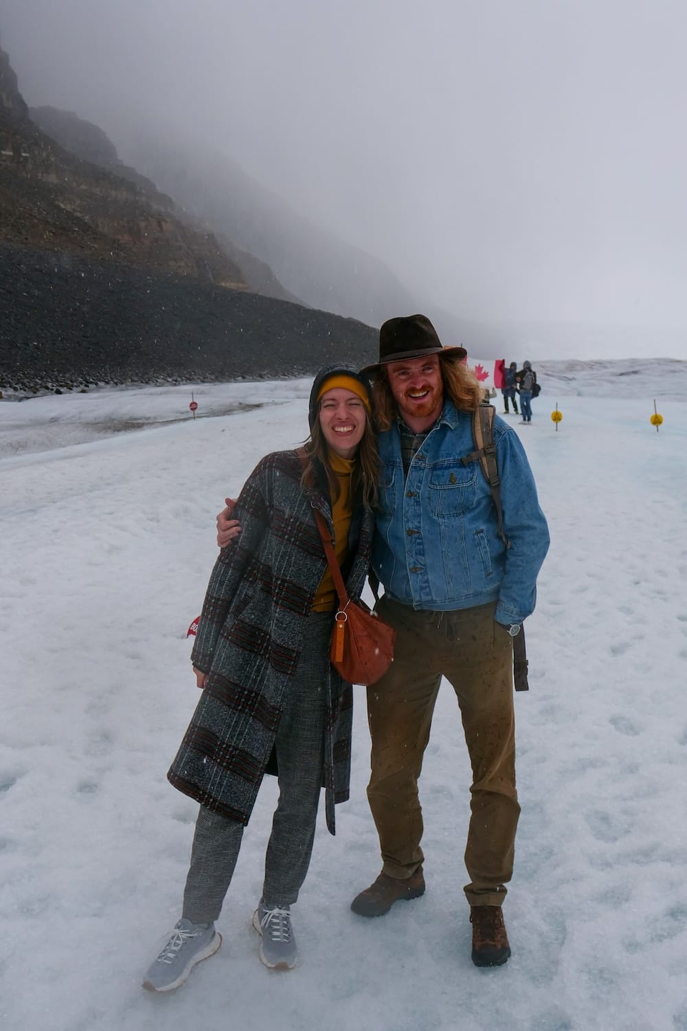 La ropa de Samuel Jeffery y Audrey Bergner en el glaciar Athabasca — Columbia Icefield, Alberta Samuel Jeffery y Audrey Bergner sonriendo juntos en el glaciar Athabasca durante una nevada, Samuel con una campera de jean y botas de trekking, Audrey con un tapado de lana a cuadros y zapatillas, Alberta, Canadá.
