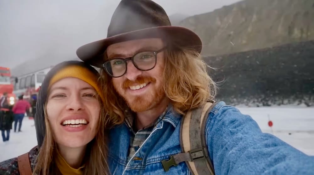 Selfie de Samuel Jeffery y Audrey Bergner en el glaciar Athabasca — Columbia Icefield, Alberta Selfie de cerca de Samuel Jeffery y Audrey Bergner sonriendo en el glaciar Athabasca mientras cae nieve y se ve un vehículo Ice Explorer detrás de ellos, Alberta, Canadá.