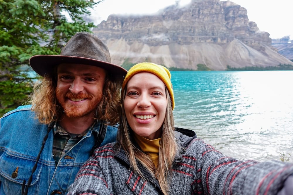 Samuel Jeffery and Audrey Bergner take a selfie together at Bow Lake along the Icefields Parkway in Banff National Park, Alberta, with the turquoise water and a mountain shrouded in low cloud behind them.