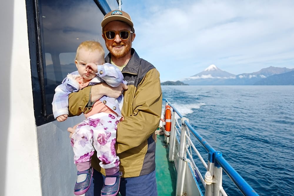 Samuel Jeffery holds Aurelia Jeffery on the deck of a boat crossing Nahuel Huapi Lake near Bariloche, Río Negro, Patagonia, Argentina, with blue water and snow-capped Andean peaks behind them.