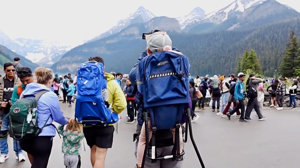 Samuel Jeffery filmando multitudes en Lake Louise con Aurelia Jeffery, Parque Nacional Banff, Alberta, Canadá. Samuel Jeffery filma la orilla llena de gente de Lake Louise en el Parque Nacional Banff, Alberta, Canadá, con la bebé Aurelia Jeffery en una mochila portabebés, rodeado por decenas de otros visitantes en el paseo de madera.