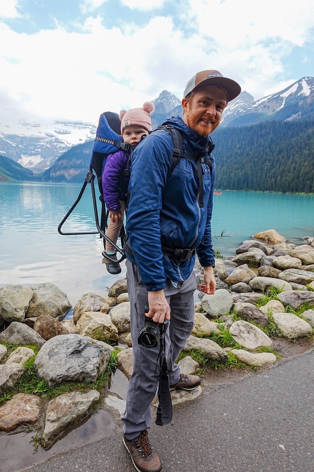 Samuel Jeffery stands on the rocky Lake Louise shoreline in Banff National Park, Alberta, Canada, with baby Aurelia Jeffery in a backpack carrier and turquoise water and mountains behind them.