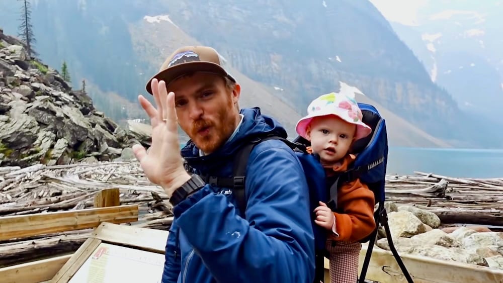 Samuel Jeffery waves at the camera on the driftwood-lined shoreline of Moraine Lake in Banff National Park, Alberta, Canada, with baby Aurelia Jeffery in a backpack carrier and turquoise water behind them.