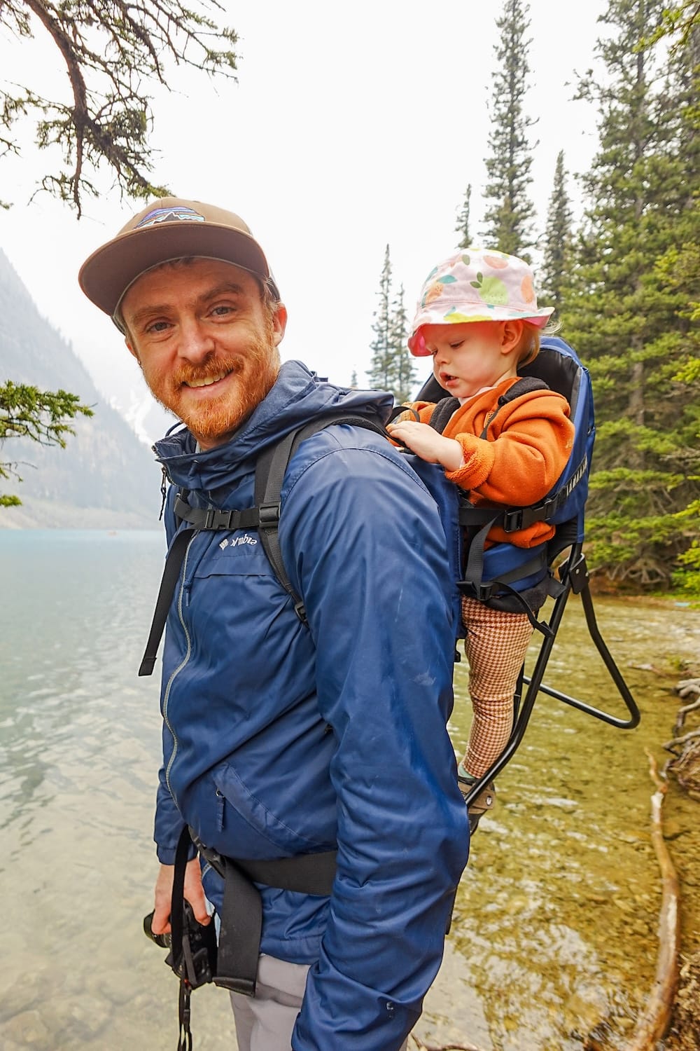 Samuel Jeffery smiles at the shallow, rocky shoreline of Moraine Lake in Banff National Park, Alberta, Canada, with baby Aurelia Jeffery in a backpack carrier and forested peaks behind them.