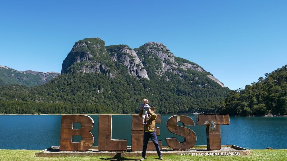 Samuel Jeffery holds his daughter Aurelia Jeffery in front of the rustic "BLEST" sign at Puerto Blest on Lago Nahuel Huapi, Patagonia, Argentina. Part of the Project 23 Patagonia Fieldwork documentation proving first-person regional expertise.